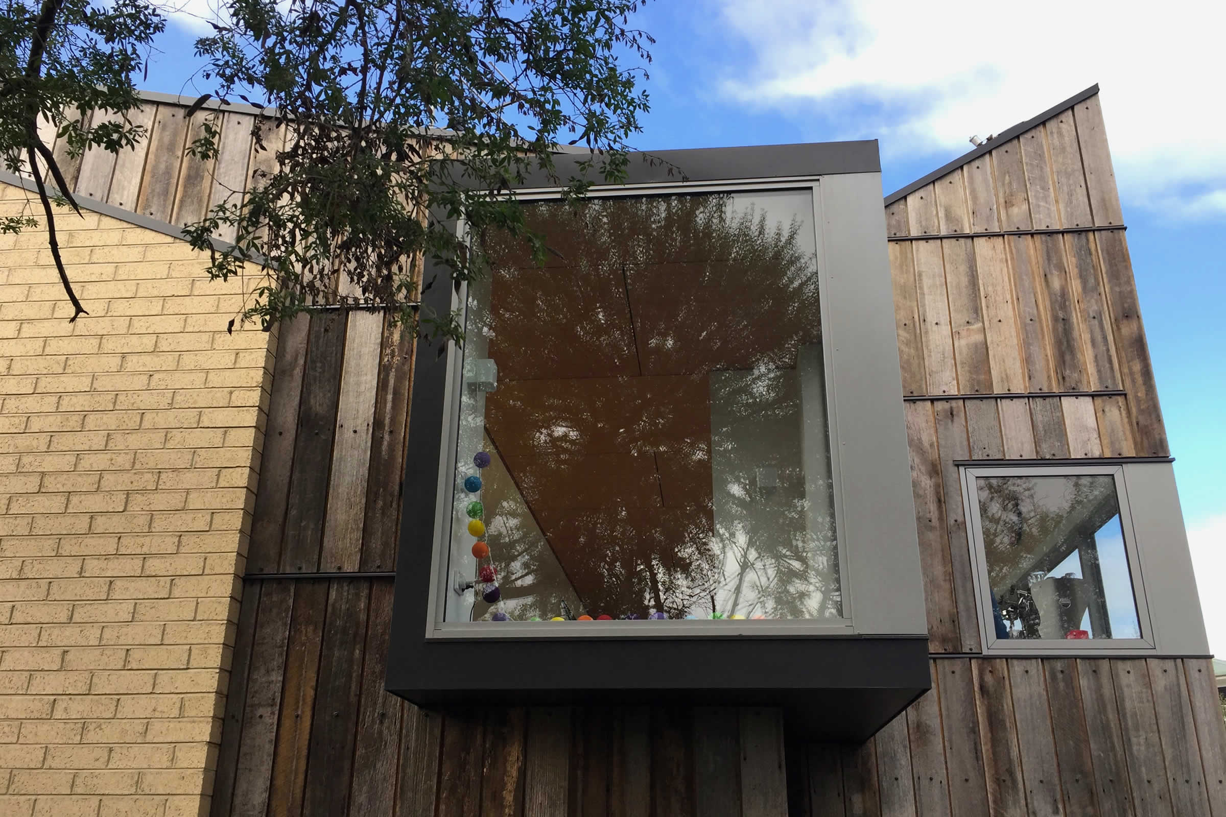 Begonia Street House, Lindisfarne, Tasmania: A corner alcove window projects beyond the plane of the distinctive recycled Tas Oak fence paling cladding and clay brick veneer exterior to capture natural light and warmth, and river and mountain views. Photo by James Morrison.