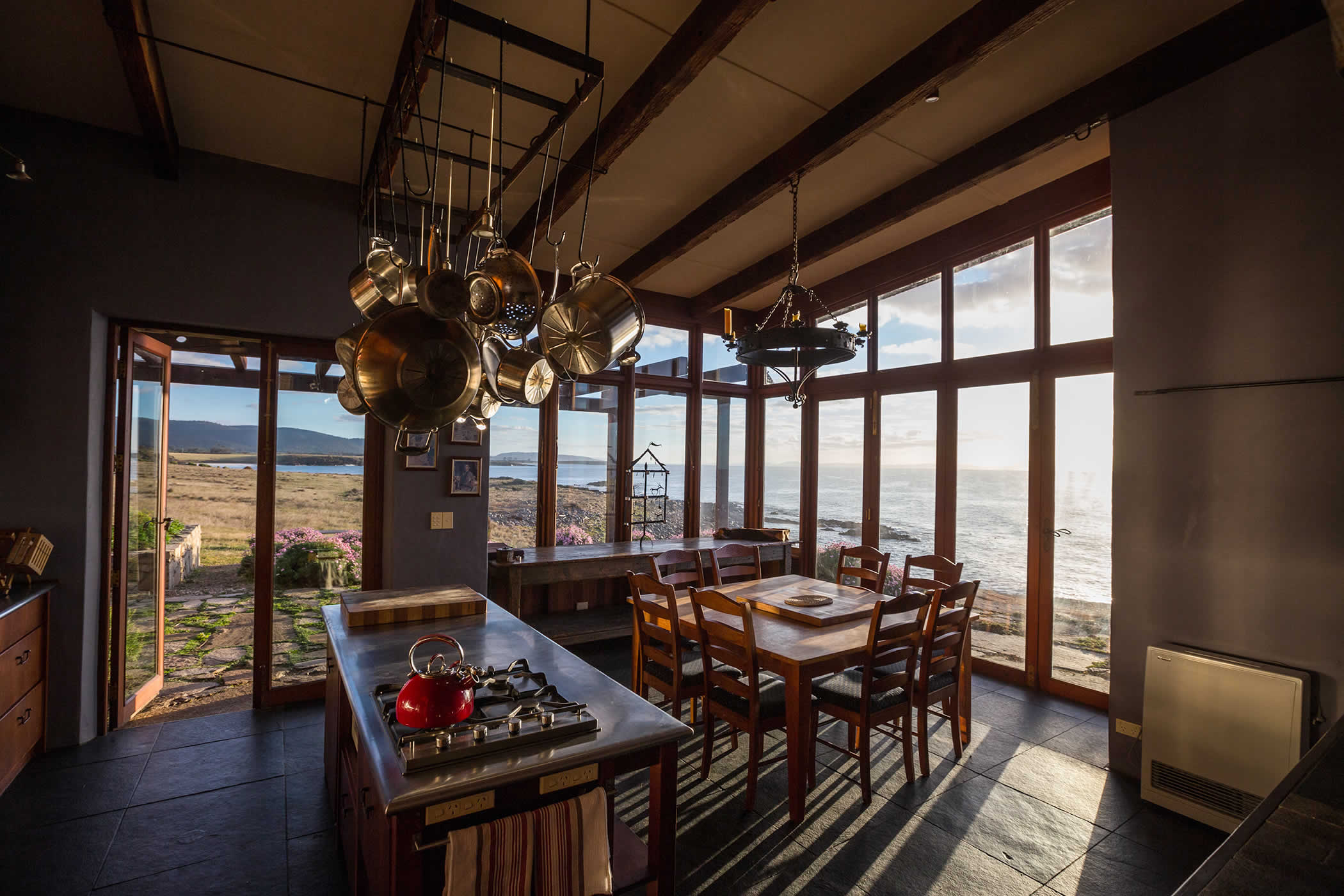 Thalia Haven, east coast Tasmania: Golden morning light washes across flagstone flooring of the luxury Thalia Haven kitchen / dining pavilion and recycled hardwood timber doors open to waterviews and private coastline. Photo by Matt Glastonbury.