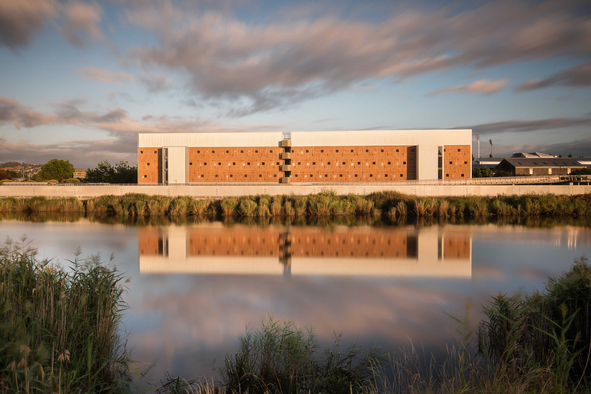 UTAS Inveresk Student Residences, Tasmania: Looking across the North Esk River to the building’s south facade and its reflection, raw macrocarpa timber screens evoke the adjacent delicate river foliage. Photo by Thomas Ryan.